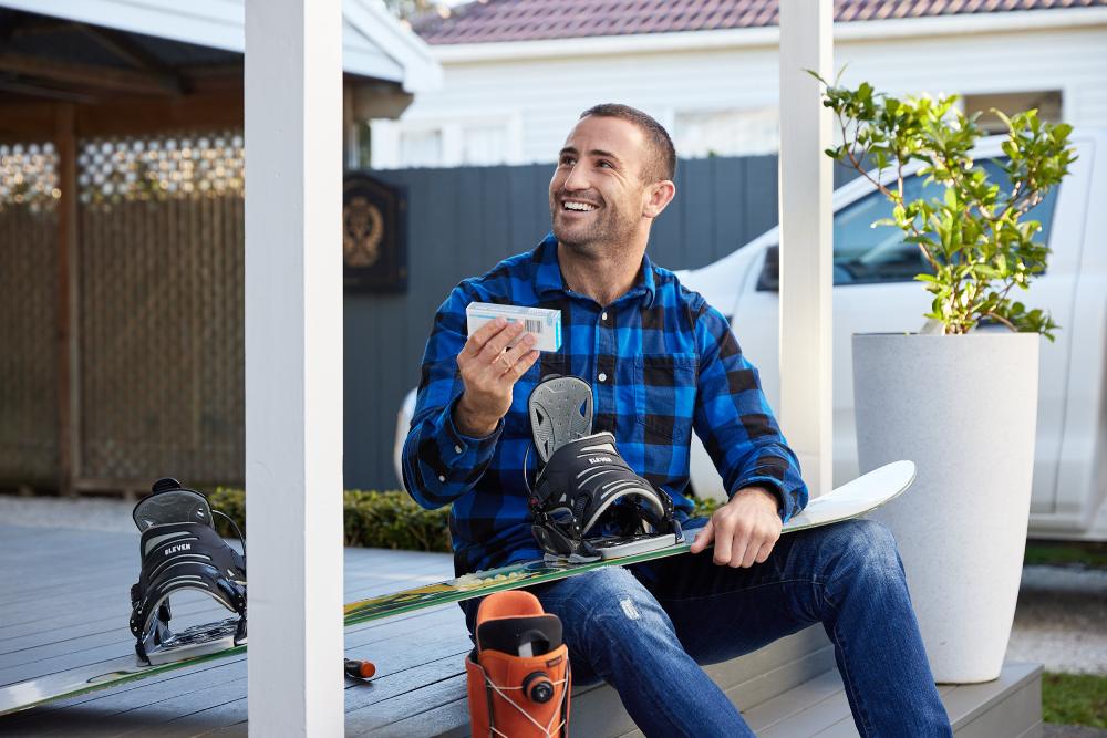 Man holding a snowboard on his lap and a Blis probiotic in his hand with a smile on his face